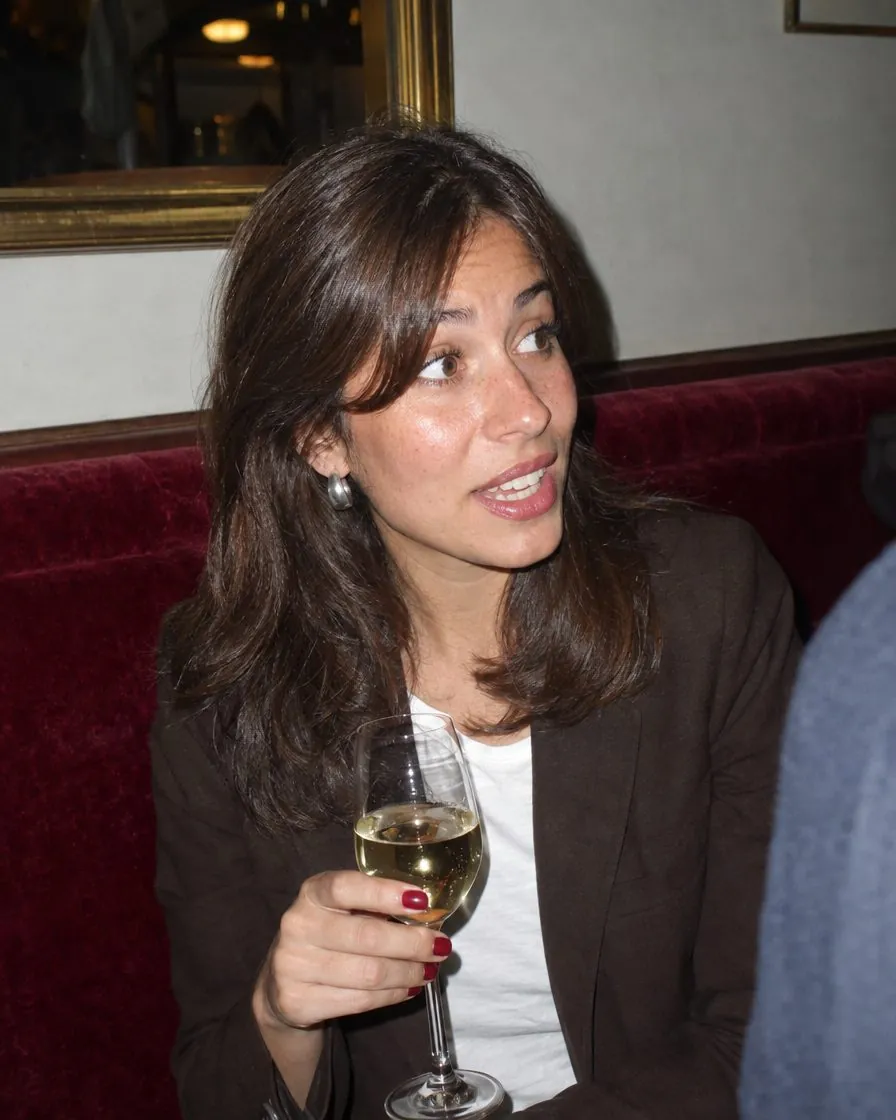 Une femme raconte quelque chose, un verre de vin blanc à la main, sur une banquette rouge de brasserie.