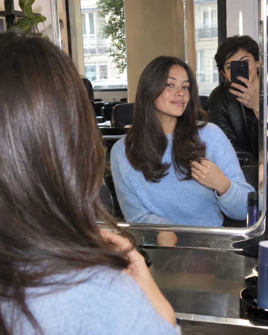 Dans un salon, une cliente fraîchement coiffée se regarde dans le miroir pendant qu'une amie la photographie.
