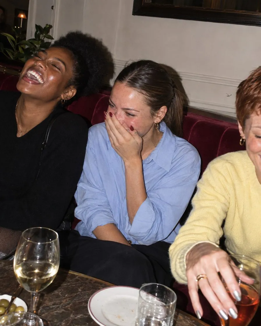 Trois amies rient sur une banquette de café, verres posés sur la table.